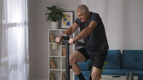 Hombre-Cansado-Está-Entrenando-Con-Bicicleta-Estática-En-Casa-Plano-Medio-De-Una-Persona-De-Pelo-Gris-Con-Bigote-Ejercicio-Cardiovascular